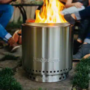 Stainless steel fire pit with flames, surrounded by people in a casual outdoor setting.