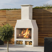 An outdoor fireplace made of volcanic pumice blocks with a visible fire burning inside, situated against a wooden fence backdrop with firewood stored underneath.